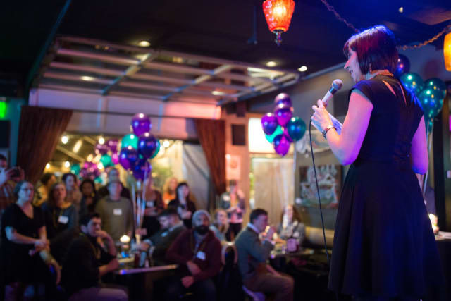 Woman speaking into a microphone at a lively event, with an audience engaged and colorful balloons in the background, capturing the atmosphere of the Love Bomb 2017 event supporting My Voice Music.